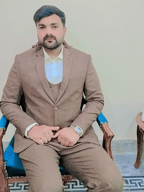 Descriptive: Portrait of a man, Mudassar Hussain, wearing a brown three-piece suit and a watch, sitting on an ornate blue chair.